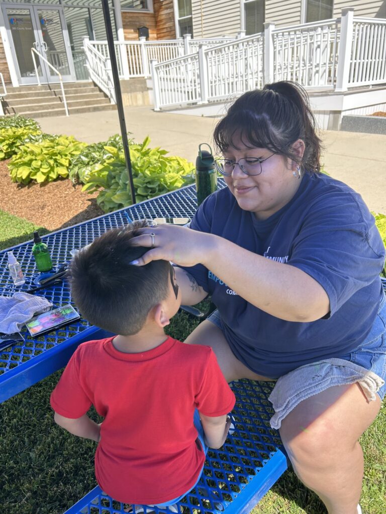 a young boy in red sits to get his face painted by a young woman in navy blue