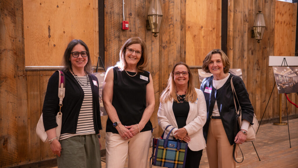 Four people pose for a photo in front of a wooden wall, one in a striped shirt with a black sweater, one in a black shirt, one in a black shirt with a while blazer and one in a white shirt with a black buttoned sweater.