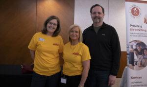 Three people pose for a photo in front of a wooden wall, with two wearing The Caring Force shirts and one wearing a black shirt.