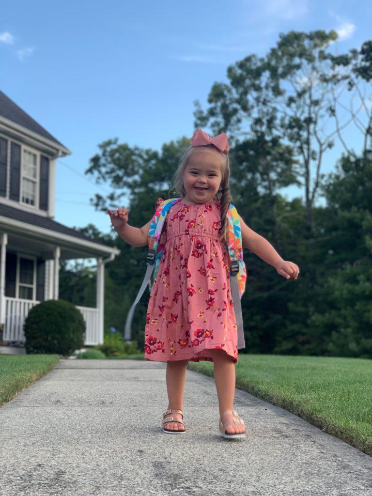 Little girl wearing a backpack walking in front of a house.